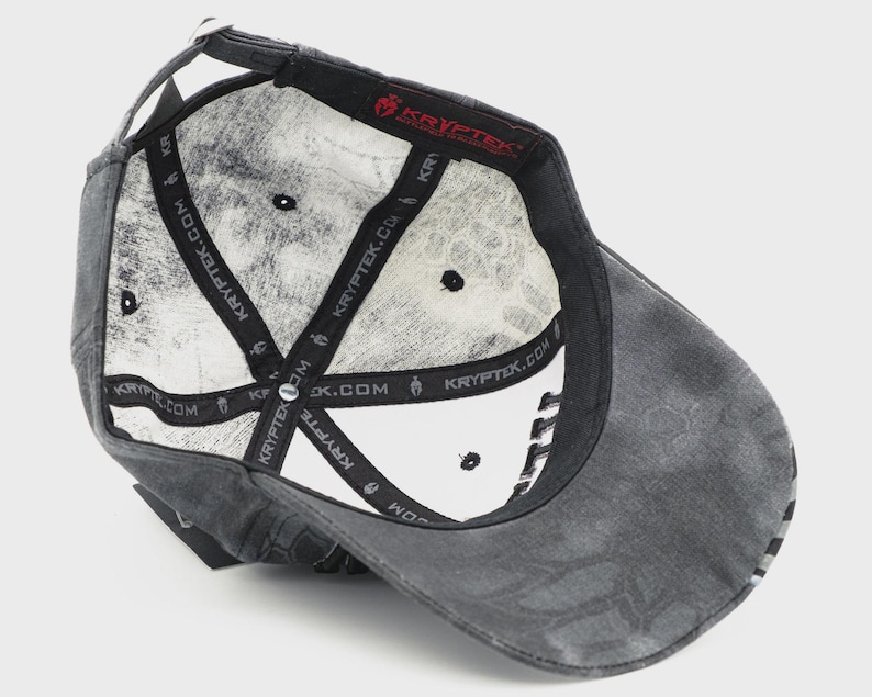 May include: A gray baseball cap with a distressed finish, viewed from the inside. The interior features a white pattern and black straps with "KRYPTEK.COM" printed on them. The cap has a curved brim and a small, black button on top.