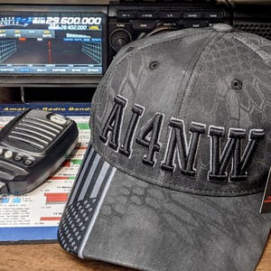 Lifestyle image of a ham radio callsign embroidered hat placed beside an amateur radio transceiver and microphone on a desk.