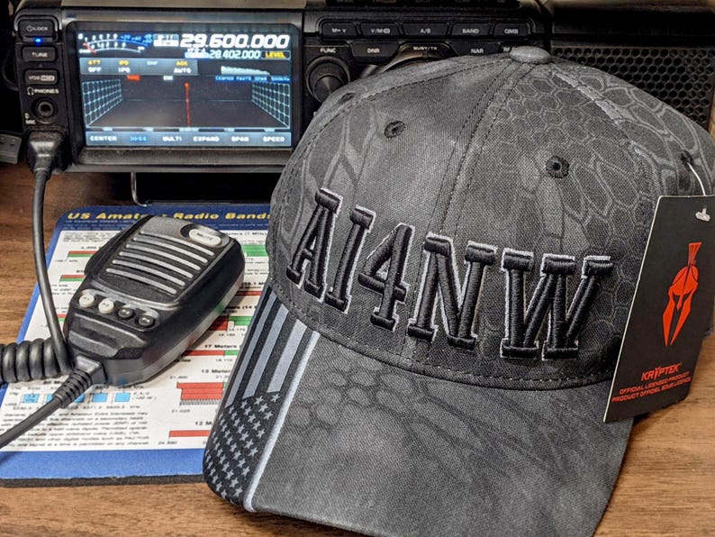 Lifestyle image of a ham radio callsign embroidered hat placed beside an amateur radio transceiver and microphone on a desk.