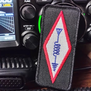 May include: Black rectangular patch with a red and white diamond design, featuring a blue antenna symbol. The patch is placed in front of a radio control panel with various buttons and a digital display.