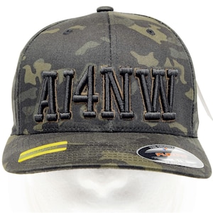 May include: A baseball cap featuring a dark green and black camouflage pattern with the text AI4NW embroidered in black on the front. The cap includes a yellow Multicam sticker on the left side of the brim and a circular Flexfit sticker on the right.