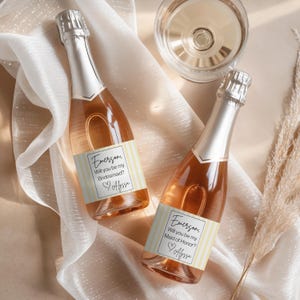 May include: Two champagne bottles with silver foil tops and labels that read "Emerson, Will you be my Bridesmaid? Alyssa" and "Emerson, Will you be my Maid of Honor? Alyssa". The bottles are on a cream-colored fabric with a glass of champagne.