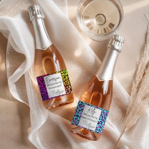 May include: Two champagne bottles with silver foil tops and labels that read "Emerson, Will you be my Bridesmaid?" and "Kelly, Will you be my Maid of Honor?" The labels have a leopard print design in bright colors.