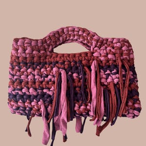May include: A crocheted handbag with a pink, red, and purple color scheme. The bag has a woven handle and a fringe detail around the bottom.