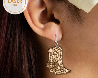 May include: Wooden laser-cut earrings featuring a cowboy boot design with sunflower accents. The earrings are light brown with intricate etched details and silver-coloured ear hooks. The earrings are hanging from an ear.