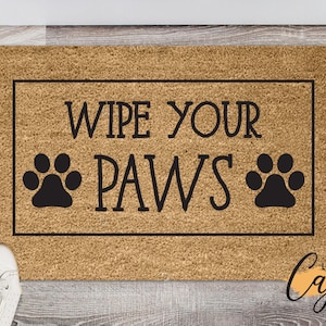 May include: A rectangular doormat made of natural fibers, featuring the text "WIPE YOUR PAWS" in black, with paw print graphics. The mat has a black border and is placed on a wooden floor. The words "Little Bit of Cajun" are in the bottom right corner.