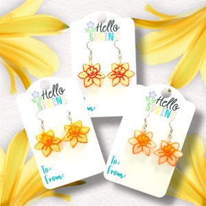 May include: Three pairs of dangle earrings featuring a yellow and orange daffodil design. Each pair is displayed on a white card with the text "Hello Spring" in colourful letters.