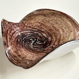 May include: A decorative glass bowl with a swirling pattern of brown and tan hues. The bowl has an asymmetrical shape with a white rim and base. The glass has a glossy finish, reflecting light. The bowl is likely for decorative use.