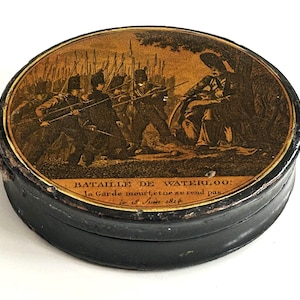 May include: A small, round, black container with a sepia-toned illustration on the lid depicting a battle scene. The text "Bataille de Waterloo" is visible. The container has a gold-coloured trim.