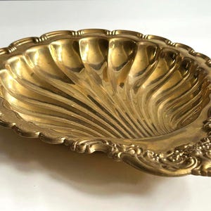 May include: A polished brass dish shaped like a scallop shell with a scalloped edge and ornate, embossed floral detailing on the handle area. The interior features deep, radiating grooves that extend from the base to the outer rim.