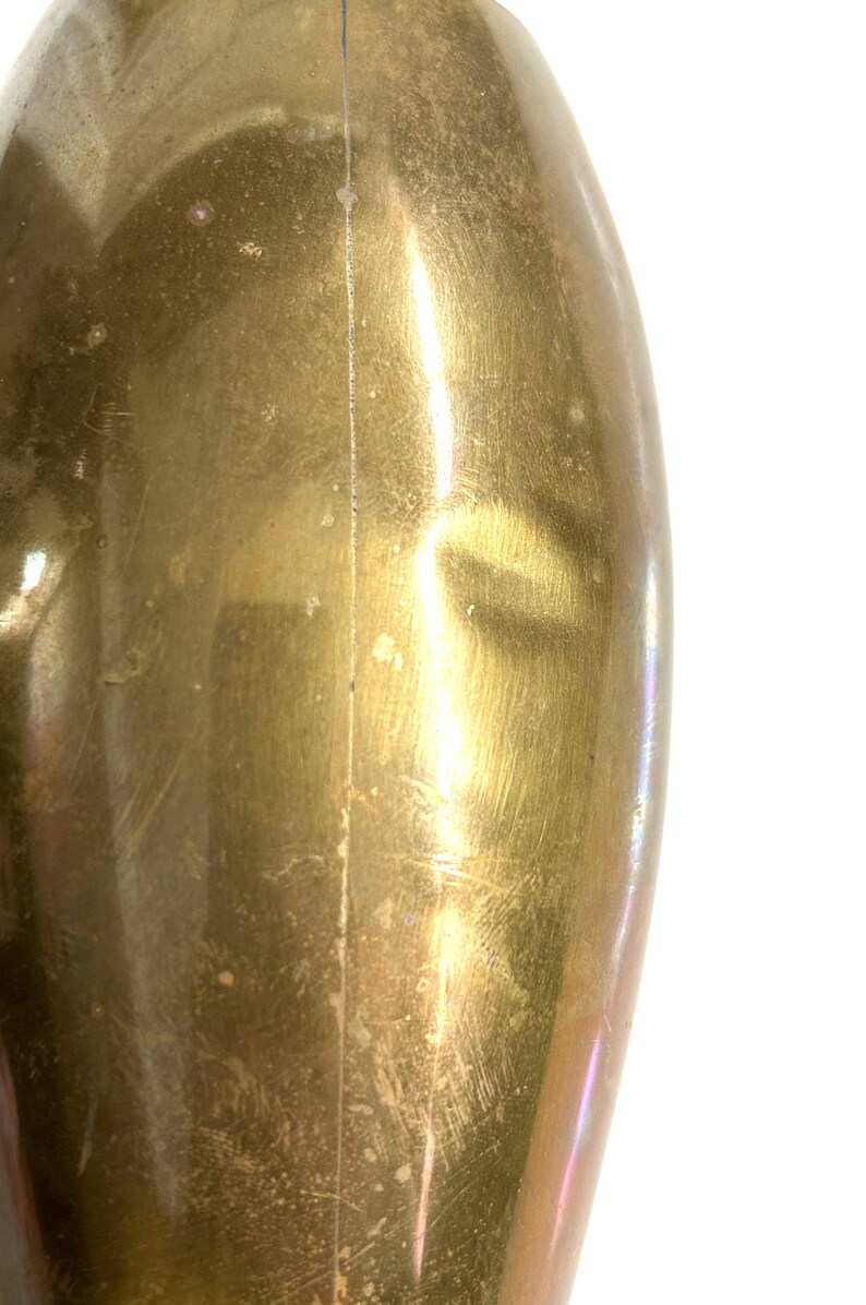 Puede incluir: Primer plano de un jarr&oacute;n de metal de forma ovalada de color dorado con una costura vertical en el centro. El jarr&oacute;n tiene una superficie lisa y pulida con algunos ara&ntilde;azos e imperfecciones.