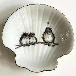 Può includere: Piatto bianco a forma di conchiglia con bordo dorato. Il piatto presenta un'illustrazione stravagante di tre uccellini appollaiati su un ramo. Gli uccelli sono raffigurati in tonalità marroni e grigie, aggiungendo un tocco di fascino naturale.