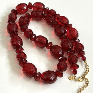 May include: A red beaded necklace with a gold-coloured clasp. The beads are faceted and of various sizes and shapes, creating a textured design. The necklace is displayed on a white surface.