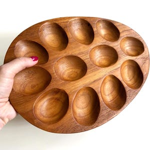 May include: An oval-shaped wooden serving tray featuring twelve individual, smooth, concave indentations arranged in a circular pattern. The tray displays a natural medium-brown wood grain texture throughout its entire surface area.