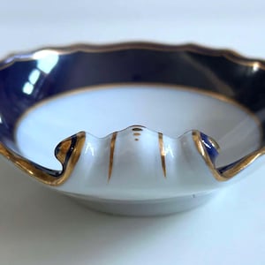 May include: A small, white porcelain dish with a blue and gold rim. The dish has a scalloped edge and a handle in the centre.