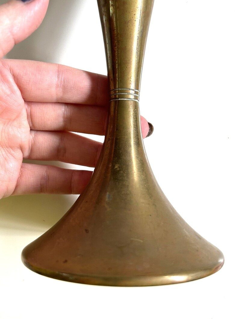 Puede incluir: Un jarr&oacute;n de metal dorado con una base acampanada y un cuello estrecho. El jarr&oacute;n tiene un acabado liso y pulido y una banda decorativa sutil alrededor del cuello.