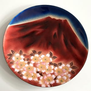 May include: A round decorative plate with a Japanese-inspired design. The plate features a red mountain with a blue sky background and a border of white and pink cherry blossoms. The plate has a glossy finish.