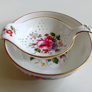 May include: A white porcelain tea strainer with a handle, featuring a colourful floral pattern of pink roses and small blossoms, resting inside a matching white porcelain bowl. Both pieces are finished with a thin gold trim along the edges.
