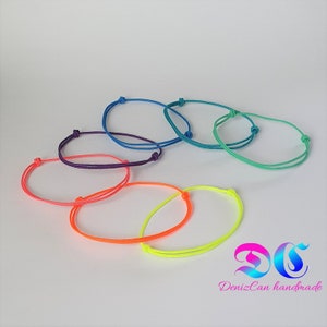 Könnte beinhalten: Ein Set aus sechs bunten Freundschaftsarmbändern aus dünnem Kordel. Die Armbänder sind in den Farben des Regenbogens, beginnend mit Lila und endend mit Grün.