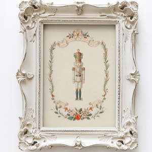 May include: A framed print featuring a central nutcracker illustration in white, gold, and blue, surrounded by a delicate botanical wreath of green leaves and red berries, enclosed in an ornate, distressed off-white vintage-style frame.