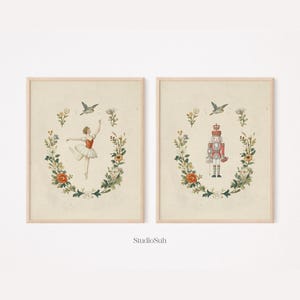 Puede incluir: Dos impresiones enmarcadas con ilustraciones vintage. Una representa a una bailarina con un corpiño rojo y tutú blanco, la otra a un soldado cascanueces. Ambas están rodeadas de coronas florales y un pájaro, con el texto "StudioSuh" en la parte inferior.