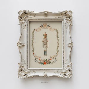 May include: A framed print of a nutcracker soldier in a white ornate frame. The print features a watercolor illustration of the nutcracker, surrounded by a wreath of greenery, berries, and flowers. The frame is distressed white.