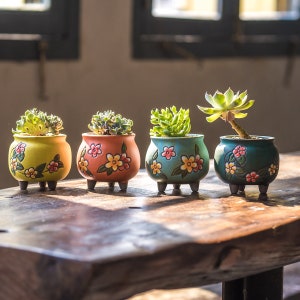 May include: Four small ceramic flower pots with painted floral designs. The pots are filled with succulents and are arranged in a row on a wooden table.