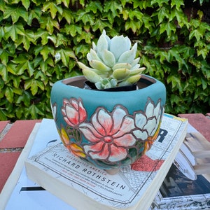 Puede incluir: Una maceta de cerámica azul verdosa con un diseño floral con flores rosas, blancas y verdes. Una planta suculenta está creciendo en la maceta.