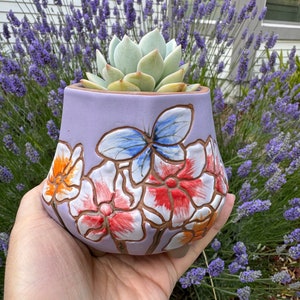 May include: A lavender ceramic planter with a floral design featuring a butterfly and flowers in shades of red, orange, and white. The planter is holding a succulent plant.