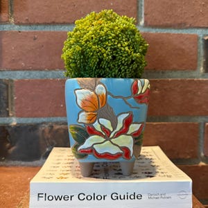 May include: A blue ceramic planter with a floral design featuring white, red, and orange flowers. The planter has three legs and is filled with a green bush.