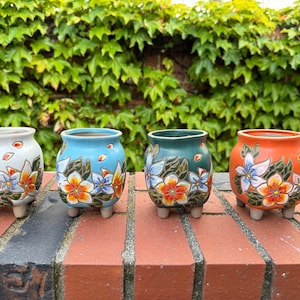 Puede incluir: Cuatro pequeñas macetas de cerámica con diseños florales. Las macetas están pintadas con flores blancas, azules, verdes y naranjas. Las macetas tienen tres patas cada una.
