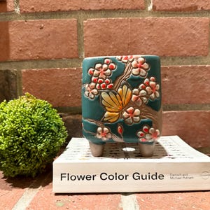 Puede incluir: Una maceta de cerámica turquesa con un diseño de mariposa y flores. La maceta tiene forma rectangular con esquinas redondeadas y se apoya en tres patas pequeñas. El diseño presenta una mariposa amarilla y flores rosas y blancas. El texto "Flower Color Guide" es visible.