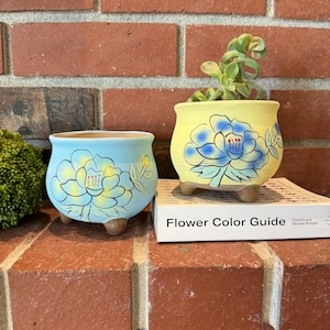 Puede incluir: Dos pequeñas macetas de cerámica con diseños florales pintados. La maceta de la izquierda es azul claro con una flor amarilla y hojas verdes. La maceta de la derecha es amarilla con una flor azul y hojas verdes. Ambas macetas tienen tres pequeños pies.