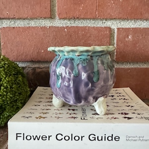 May include: A small, handcrafted ceramic pot with a rounded body and three small feet. The pot is a deep purple color with turquoise and teal glaze dripping down the sides. The pot sits on a book titled "Flower Color Guide".