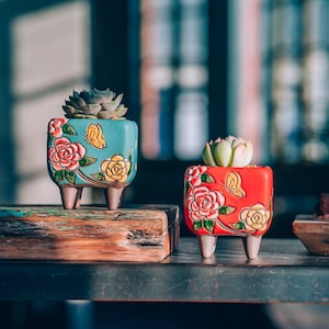Puede incluir: Dos pequeñas macetas de cerámica con diseños florales y detalles de mariposas. La maceta de la izquierda es azul con flores amarillas y una mariposa, mientras que la maceta de la derecha es roja con flores rosas y una mariposa. Ambas macetas tienen patas cortas.