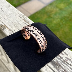 May include: A copper bracelet with a woven design. The bracelet is on a black velvet pouch.