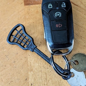 Pode incluir: Um chaveiro de taco de lacrosse preto preso a um chaveiro de carro preto com um botão prateado. O chaveiro está preso a um chaveiro prateado com uma chave de latão e uma chave prateada.