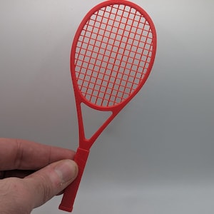 Può includere: Una racchetta da tennis rosso brillante con una griglia di corde. La racchetta è tenuta da una mano su uno sfondo bianco. L'impugnatura è strutturata.