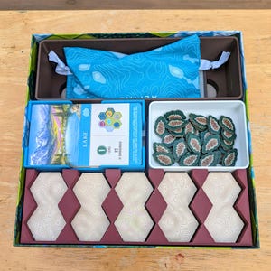 May include: A board game box containing a blue drawstring bag with topographic patterns, a deck of Lake cards, a white tray filled with small green pinecone tokens, and five light-coloured hexagonal tiles with topographic line designs.