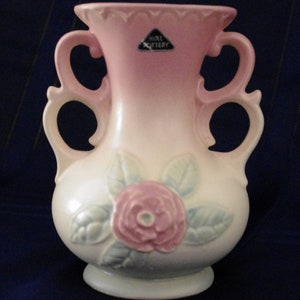 Puede incluir: Un jarrón de cerámica esmaltada en rosa y blanco con dos asas y un diseño de rosa. El jarrón está marcado con "Hull Pottery".