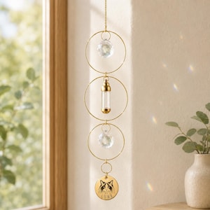 May include: A gold-toned hanging mobile featuring three thin circular hoops connected vertically. The top and middle hoops contain clear faceted crystal spheres, while the center hoop holds a small glass vial. A round gold charm with a cat illustration and the text LUNA hangs at the bottom.