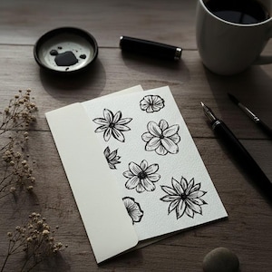 May include: A still-life image featuring a white card with black floral illustrations, nestled in a white envelope. A cup of coffee, inkwell, pen, and brush are on a wooden surface. The scene is lit with soft, natural light.