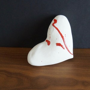 May include: A white ceramic heart-shaped ornament with red paint splatters and streaks.