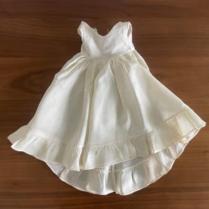 Puede incluir: Vestido de muñeca de color marfil con dobladillo alto-bajo y ribete con volantes. El vestido presenta un diseño sin mangas y un corpiño ajustado. La falda se ensancha desde la cintura, creando una silueta completa y fluida. El vestido está hecho de tela suave y ligera.