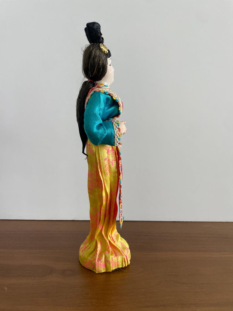 Op de afbeelding: Een decoratieve pop met donkerbruin haar in een knotje met een zwart accessoire. De pop draagt een blauwgroene top met lange mouwen en ingewikkelde randen en een lange gele rok versierd met een roze bloemenpatroon, gezien in zijaanzicht.