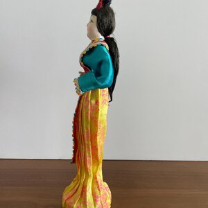 Op de afbeelding: Een decoratieve pop met donker haar in een opgestoken kapsel, gekleed in een groenblauw shirt met lange mouwen en kleurrijke randen, en een gele rok tot op de grond met een roze bloemenpatroon. De pop is afgebeeld in zijaanzicht.