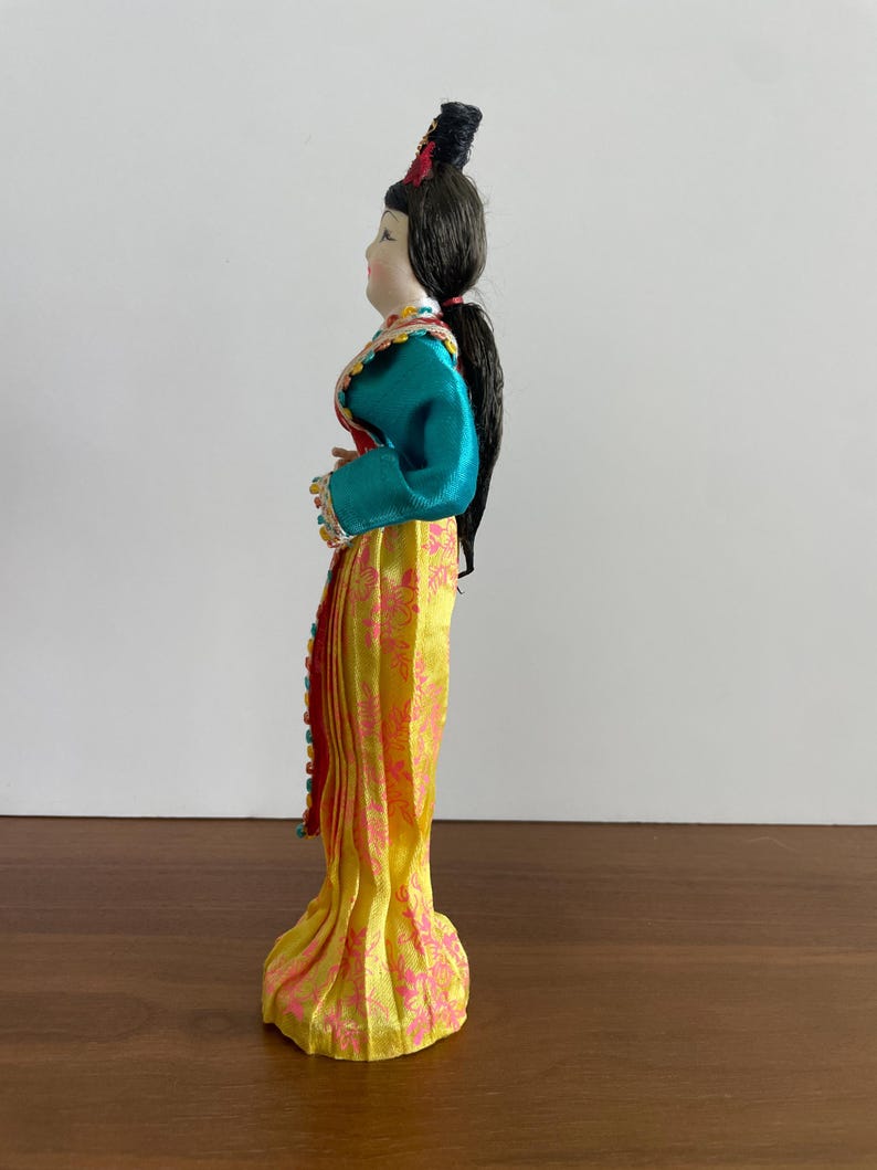 Op de afbeelding: Een decoratieve pop met donker haar in een opgestoken kapsel, gekleed in een groenblauw shirt met lange mouwen en kleurrijke randen, en een gele rok tot op de grond met een roze bloemenpatroon. De pop is afgebeeld in zijaanzicht.