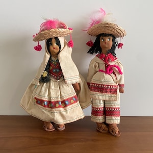 May include: Two traditional dolls in regional dress. They wear straw hats with pink feathers and tassels. One doll is in a cream dress with red and blue embroidery. The other doll is in a cream outfit with red detailing, including trousers.