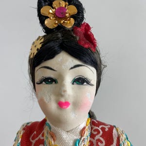 Op de afbeelding: Een pop met zwart haar in een opgestoken kapsel, versierd met een goudkleurige bloem en een rood stoffen accent. De pop heeft geschilderde gelaatstrekken, waaronder groene ogen en roze lippen, en draagt een rood en blauwgroen kledingstuk met patroon.