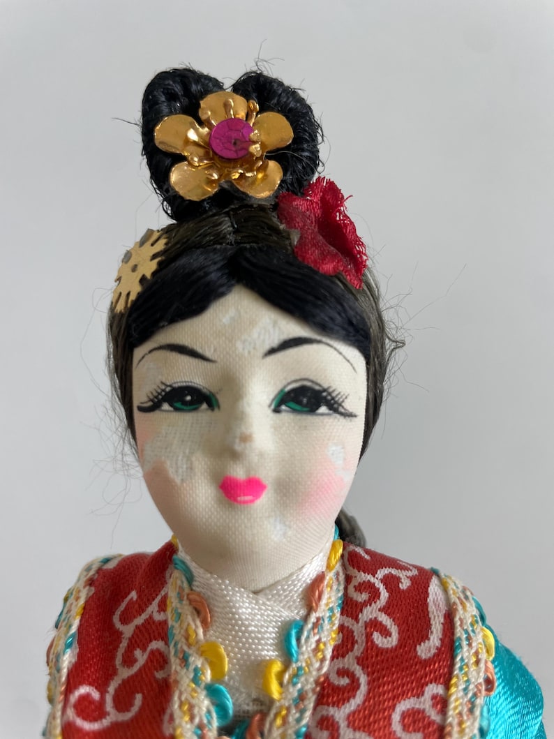 Op de afbeelding: Een pop met zwart haar in een opgestoken kapsel, versierd met een goudkleurige bloem en een rood stoffen accent. De pop heeft geschilderde gelaatstrekken, waaronder groene ogen en roze lippen, en draagt een rood en blauwgroen kledingstuk met patroon.